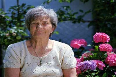 زنی سالخورده با موهای کوتاه خاکستری و لباسی سفید رنگ در حالی که کنار بوته‌ای از گلهای صورتی رنگ نشسته است